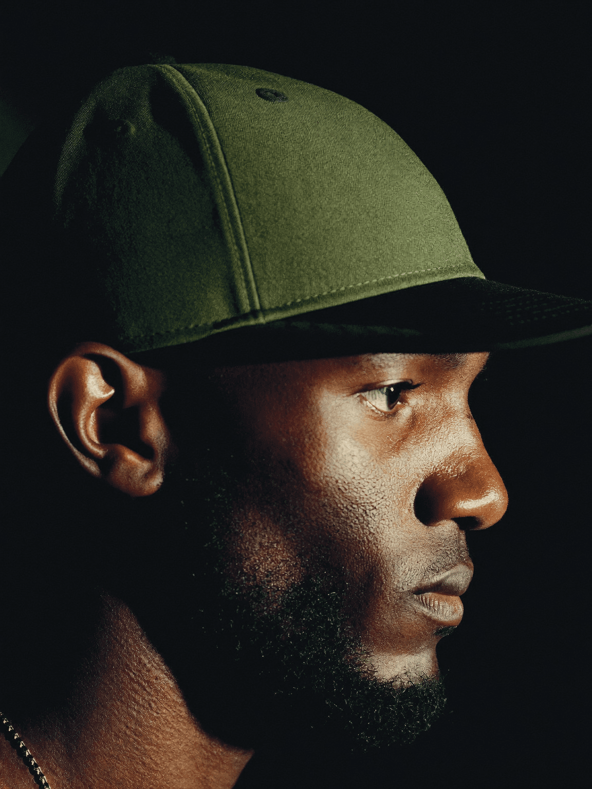 Close-up profile of a man in an olive snapback under dramatic light