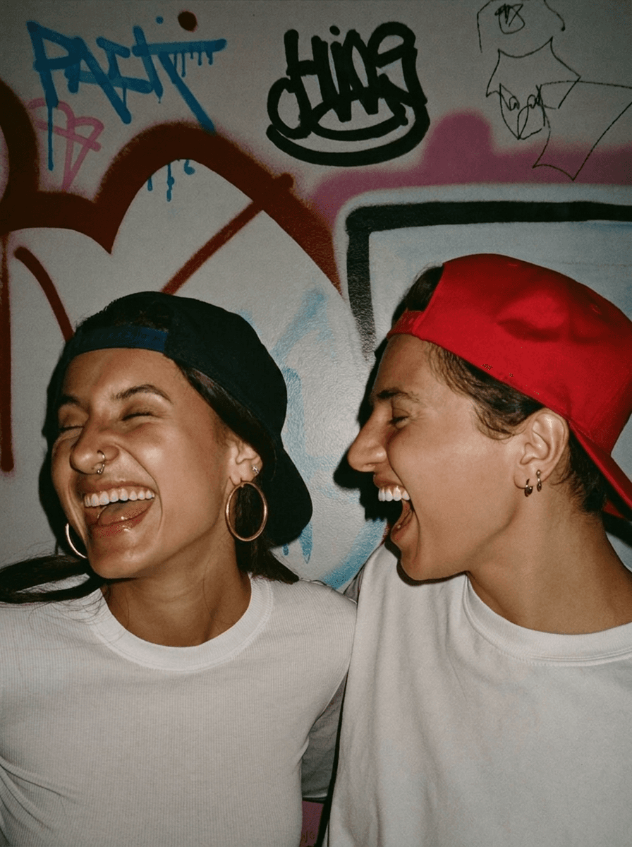Two friends laughing against a graffiti wall wearing snapbacks